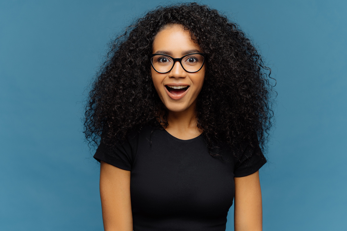 Pleasantly Surprised Dark Skinned Woman Keeps Jaw Dropped, Gazes with Interest, Has Curly Hair, Wears Black T Shirt, Models against Blue Wall. Afro American Lady Gazes with Shock, Has Bated Breath